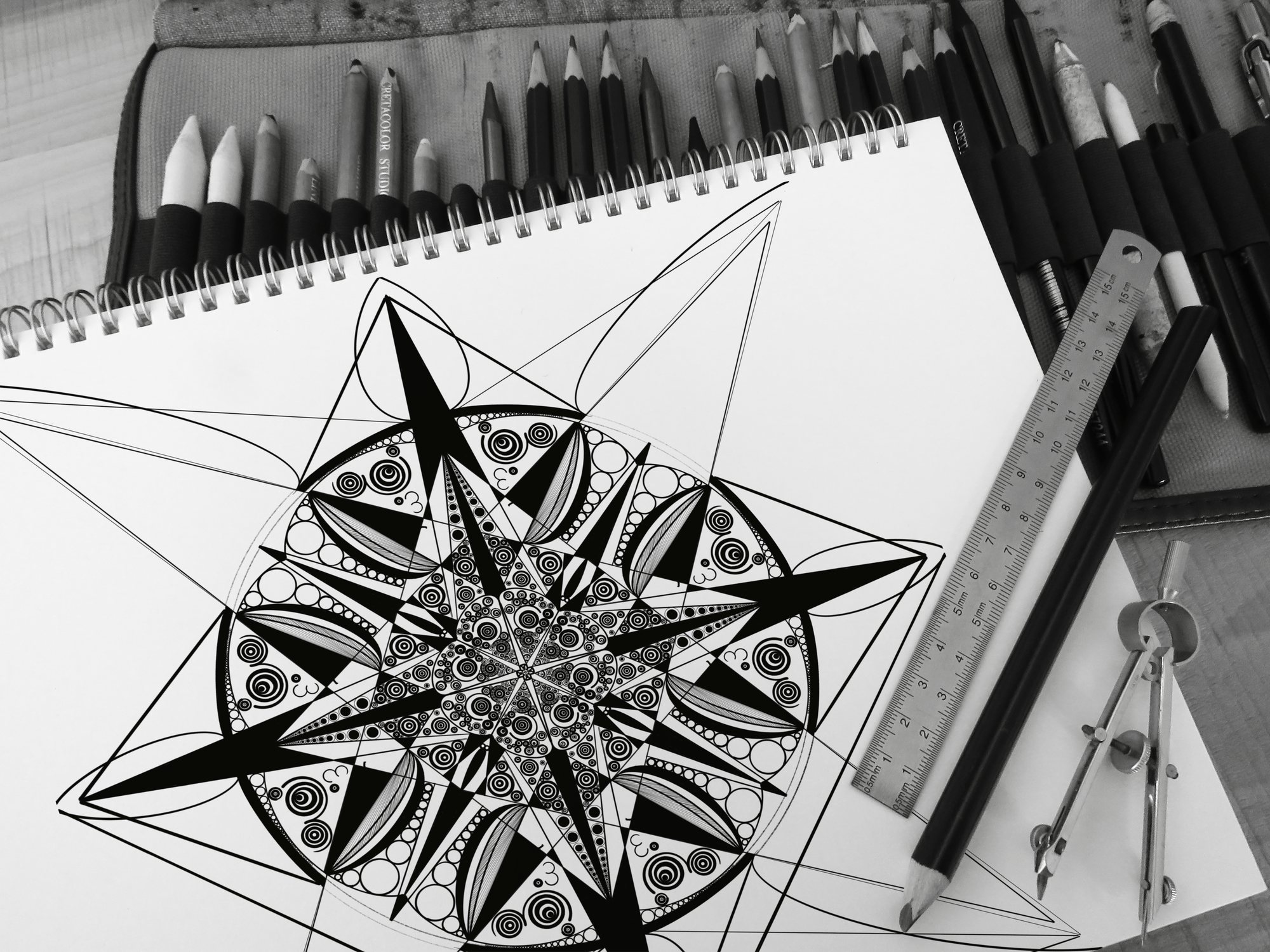 Geometric ink mandala on a sketchpad beside pencils, a ruler and a compass